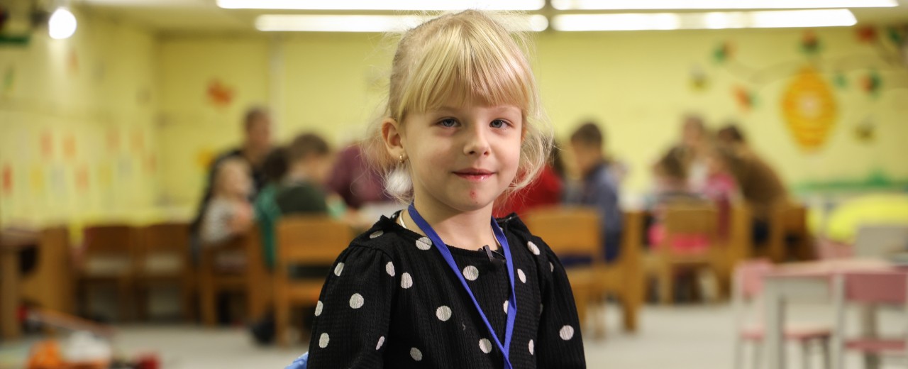 Blonde girl with a ponytail and fringes looks into the camera. Children play at a table in the background.