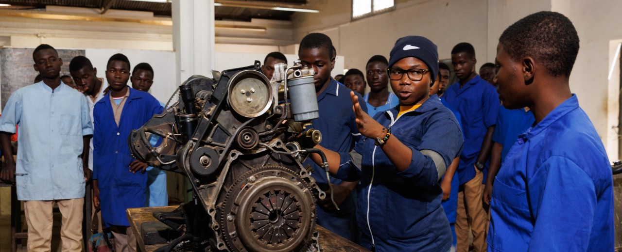 Person stands next to dismantled car engine and teaches others. 