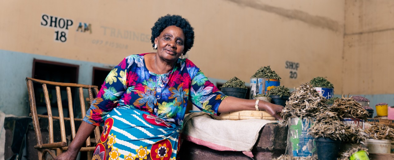 A woman is standing next to her goods, stacked on a table.