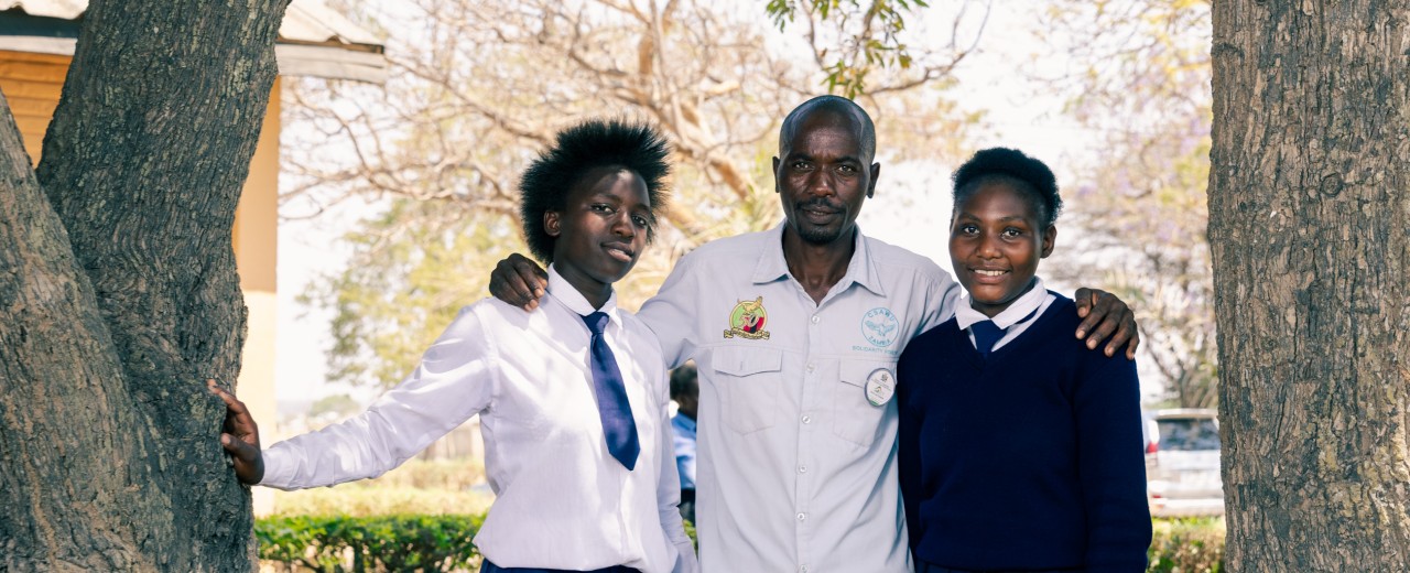 Two girls in school uniforms and their father in their middle are standing in between trees.