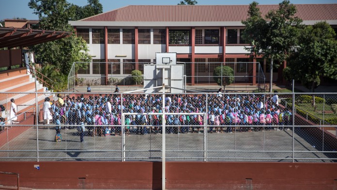 Primary school in Maputo: three teachers gather the pupils on the school's sports field with the school building in the background.