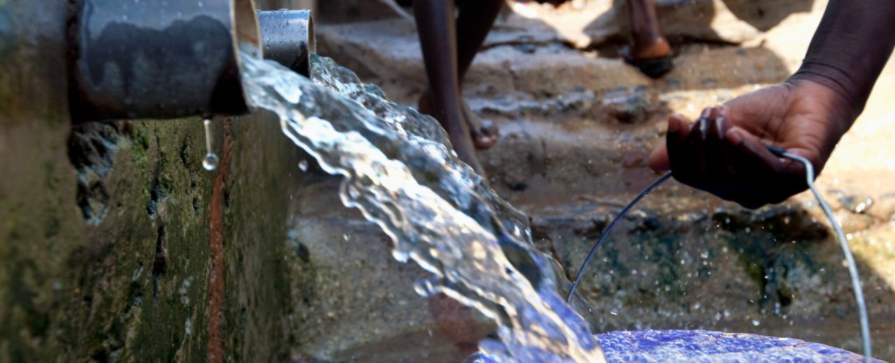 Aus einem Trinkwasserbrunnen fließt Wasser in einen blauen Plastikeimer.