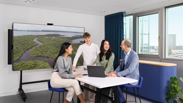 Eine diverse Gruppe (Personen verschiedenen Geschlechts, Alters und Herkunft) sitzt zusammen an einem Besprechungstisch und diskutiert angeregt über nachhaltige Waldwirtschaft. Im Hintergrund ist auf einem großen Monitor ein Bild vom Regenwald zu sehen.