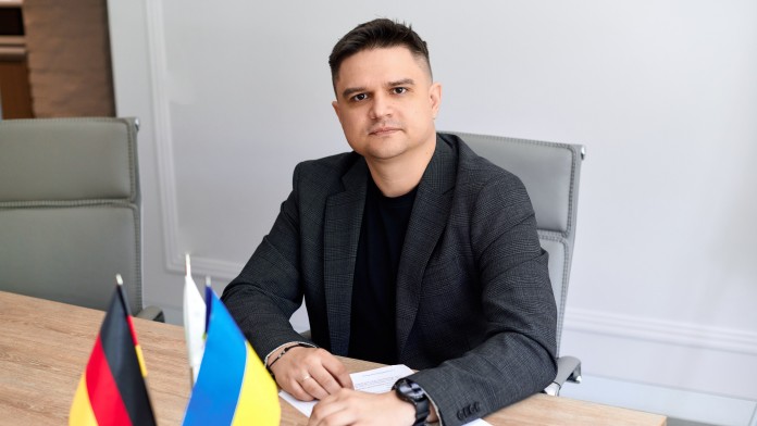 A man in a suit is sitting at a desk looking into the camera. In front of him on the table are a German and a Ukrainian flag.