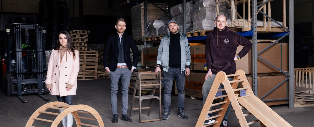 A young woman and three young men are standing in a warehouse. They have wooden play equipment for children in front of them.