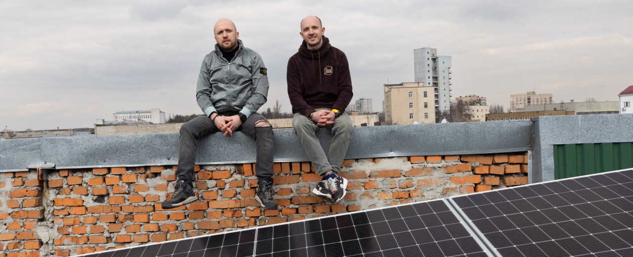 Two young men sit on a wall above the rooftops. Solar panels can be seen at their feet.