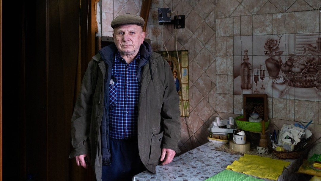 An older man in a cap and thick jacket is standing at a table in a kitchen tiled to the ceiling. Above the table is a tiled picture.