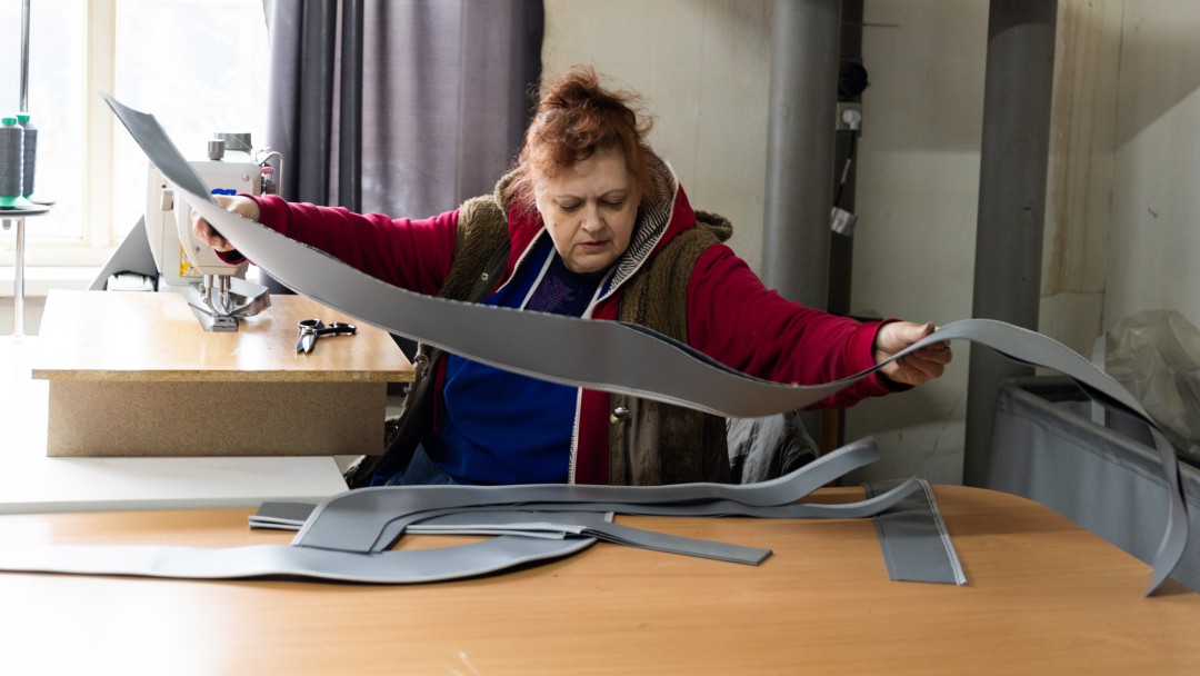 A woman spreads out cut leather parts on a table. A sewing machine can be seen in the background.
