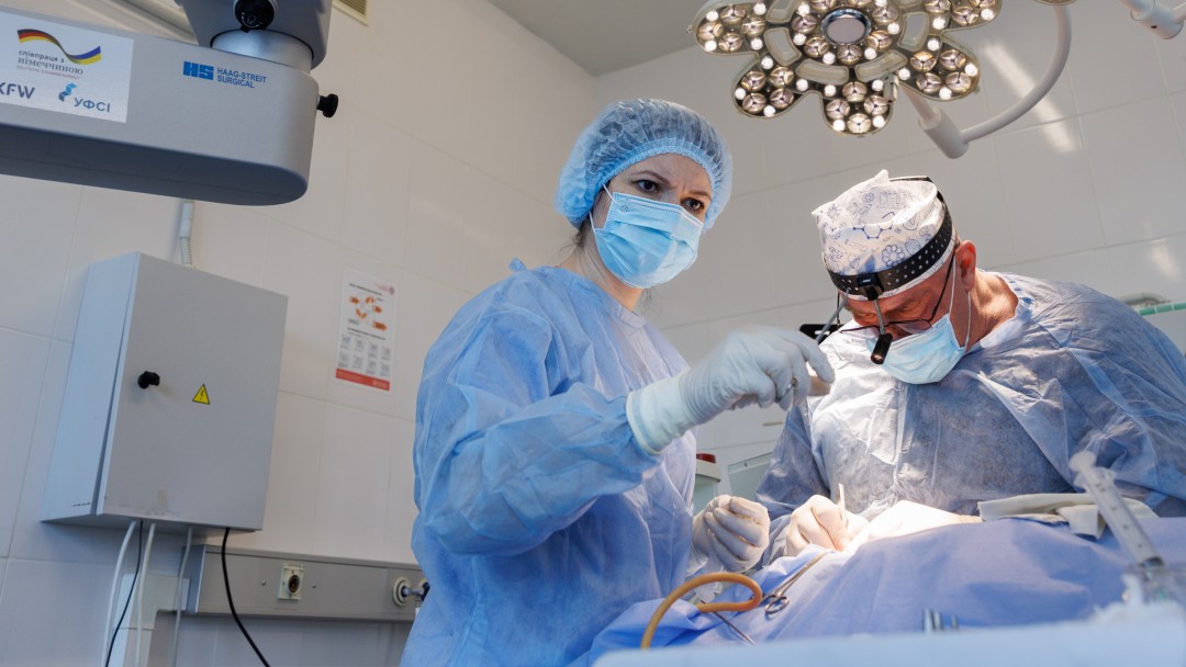 Two doctors in blue surgical gowns bend over the operating table. A technical device can be seen in the foreground.