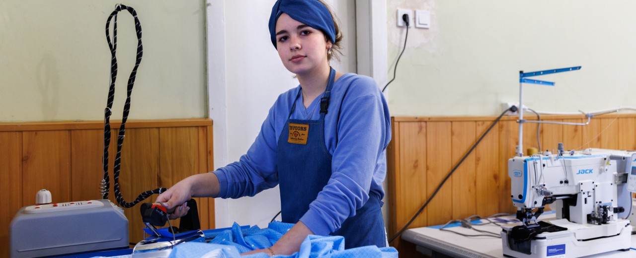 Eine junge Frau bügelt an einer Bügelstation blauen Stoff. Im Hintergrund ist eine Nähmachine zu sehen.