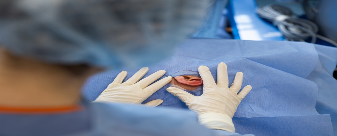A pair of hands wearing rubber gloves hold a blue surgical drape over a patient's head. There is a hole in the cloth through which an ear can be seen.